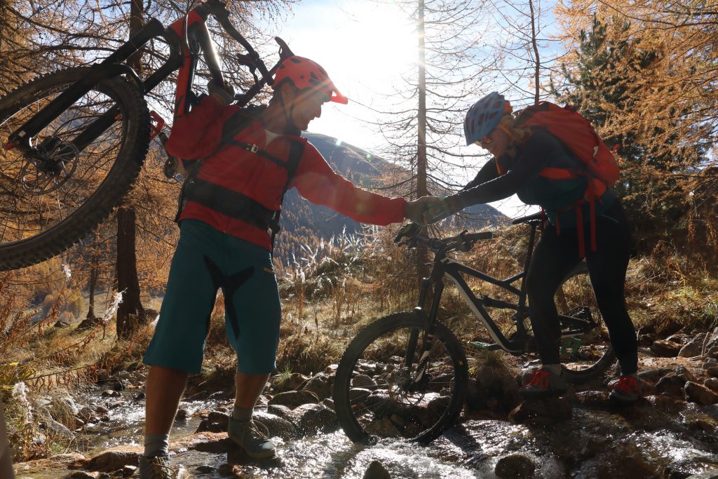 Mountain bikers cross mountain creek, man offers assistance.